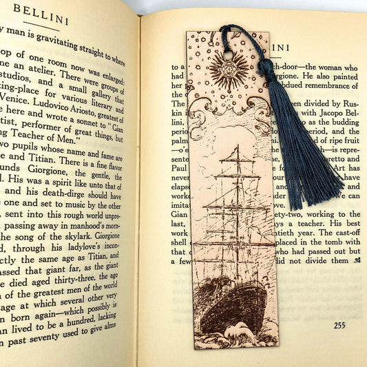 A leather bookmark with the historical art of a ship engraved onto it. This bookmark has a dark blue tassel and is resting on the pages of an open book. Leather Ship Bookmark Bookmarks Raven King Crafts