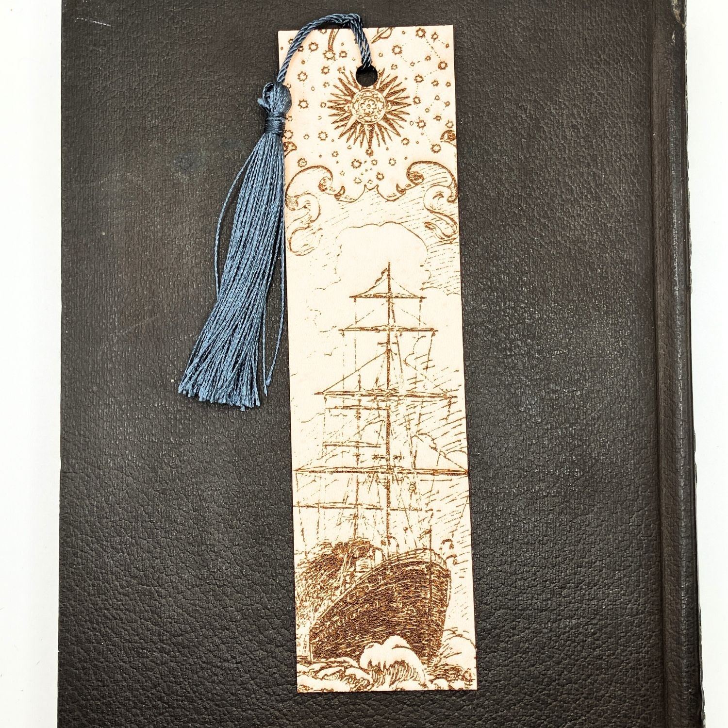 A leather bookmark with the historical art of a ship engraved onto it. This bookmark has a dark blue tassel and is resting on the pages of a closed leather book.