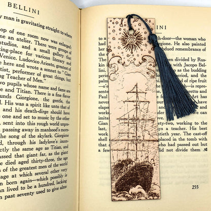 A leather bookmark with the historical art of a ship engraved onto it. This bookmark has a dark blue tassel and is resting on the pages of an open book.