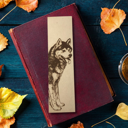 Photograph of the husky bookmark sitting on top of a closed red covered book. The book is resting on a dark wood table with orange leaves around it and part of a tea cup visible.