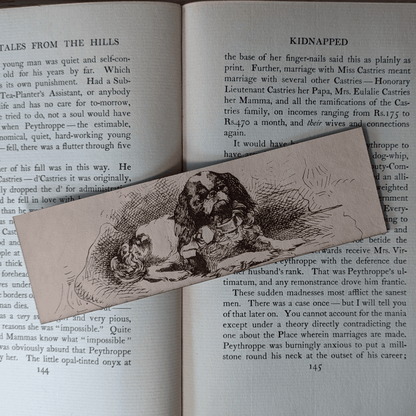 A rectangular leather bookmark with the historical art of a small spaniel dog engraved on it. The bookmark is resting on the pages of an open book. Leather Spaniel Bookmark Bookmarks Raven King Crafts Leather Spaniel Bookmark Bookmarks Raven King Crafts