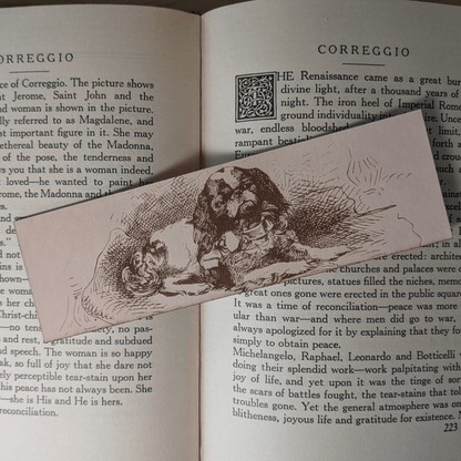 A rectangular leather bookmark with the historical art of a small spaniel dog engraved on it. The bookmark is resting on the pages of an open book. Leather Spaniel Bookmark Bookmarks Raven King Crafts Leather Spaniel Bookmark Bookmarks Raven King Crafts
