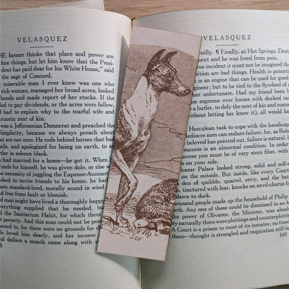 A rectangular leather bookmark with the historical art of a Whippet dog engraved on it. The bookmark is resting on the pages of an open book. Leather Whippet Dog Bookmark Bookmarks Raven King Crafts Leather Whippet Dog Bookmark Bookmarks Raven King Crafts