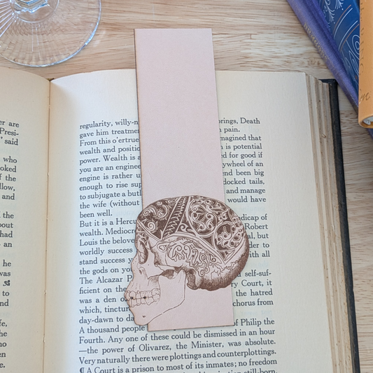 A leather bookmark engraved with the art of a skull on it. The skull bookmark is resting on the pages of an open book. 