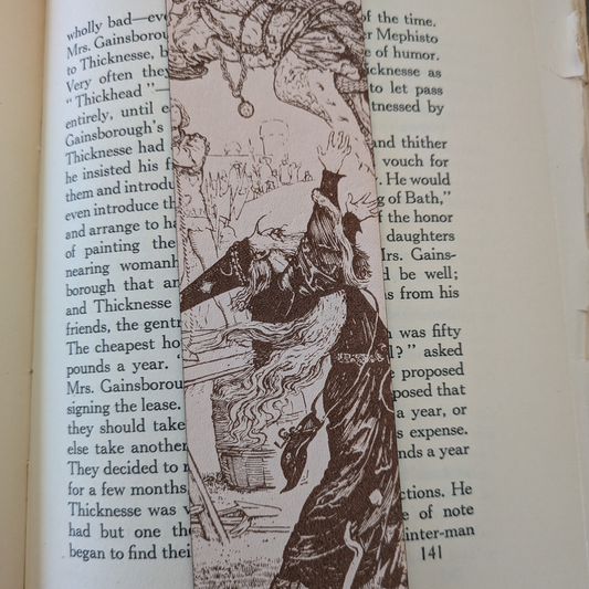 A leather bookmark engraved with the art of a wizard on it. The wizard bookmark is resting on the pages of an open book. 