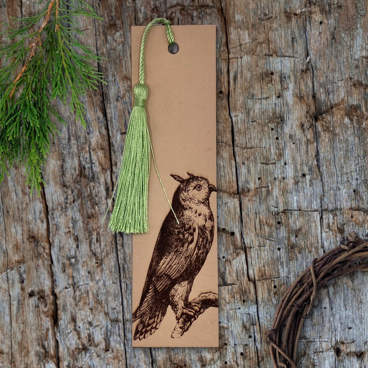 A leather bookmark with the art of a long - eared owl engraved on it. The leather bookmark has a green tassel. Next to the bookmark is a evergreen twig and a small wooden wreath.