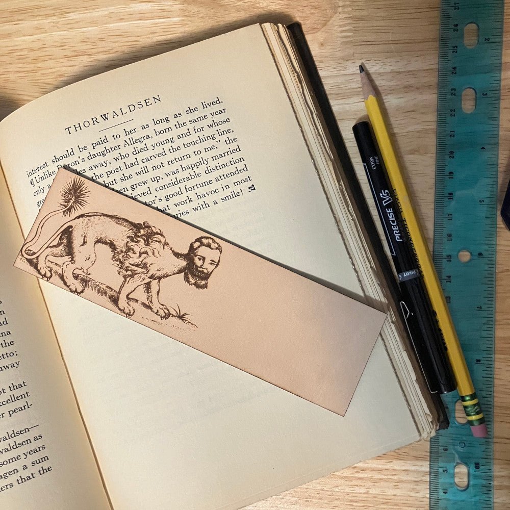 A leather bookmark with an illustration of a manticore engraved onto it. The bookmark is resting on the pages of an open book.