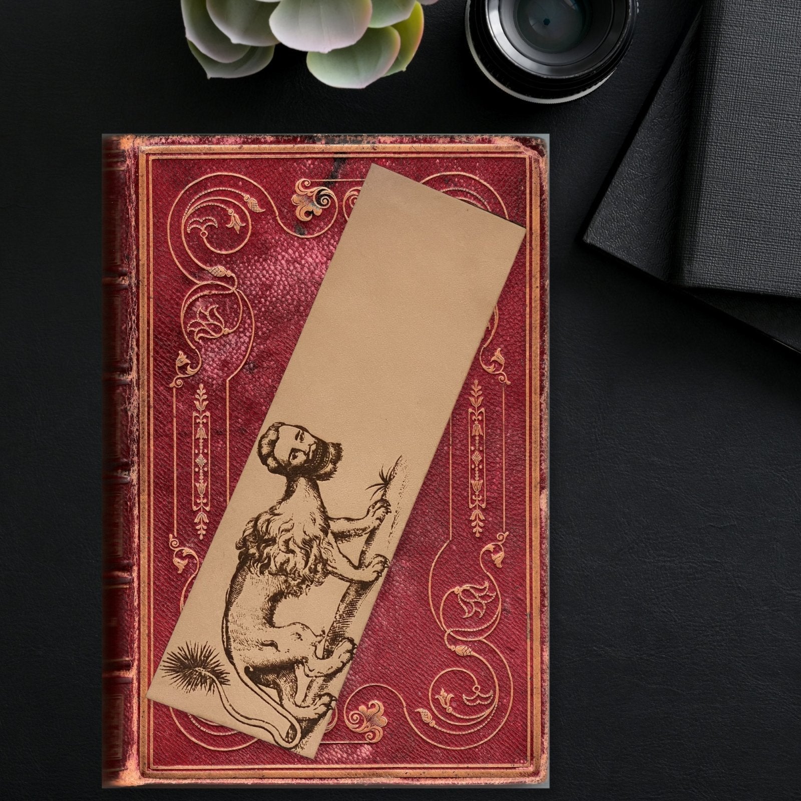 A leather bookmark with the art of a manticore on it. The creature has the body of a lion, a spiked tail and the head of a man with sharp teeth. The bookmark is resting on a red book.