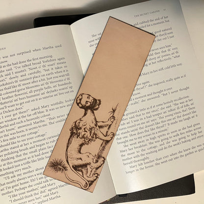 A leather bookmark with an illustration of a manticore engraved onto it. The bookmark is resting on the pages of an open book.