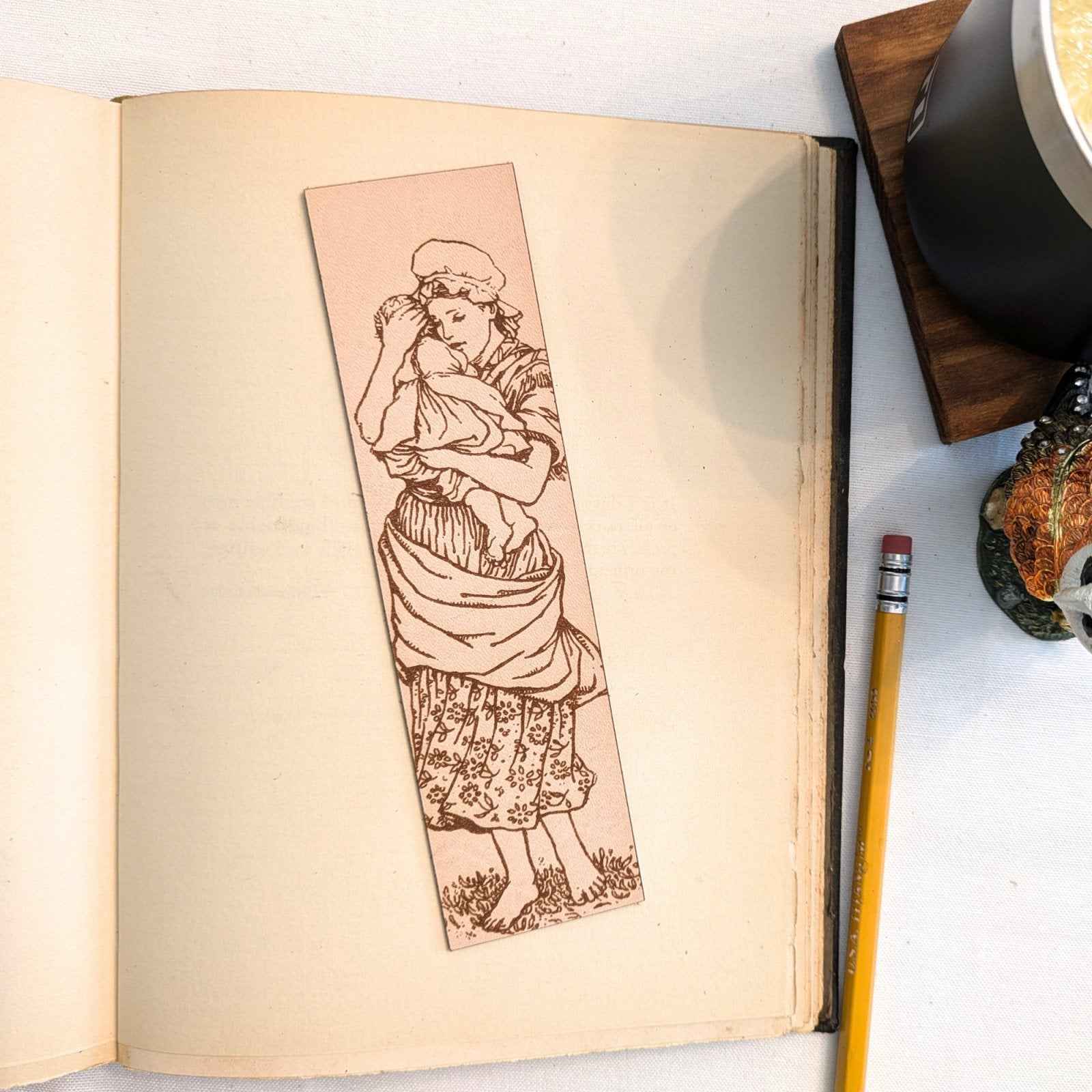 A photograph of historical mother holding a child leather bookmark resting on the pages of an open book. Mother and Child Leather Bookmark Bookmarks Raven King Crafts