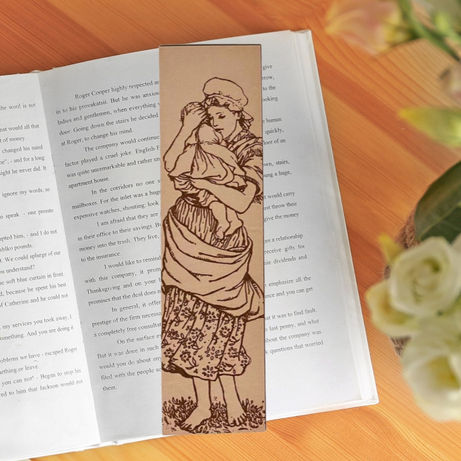 A photograph of a bookmark resting on the pages of an open book. With flowers out of focus. The bookmark is a rectangle in shape with the art of a mother holding her child. They are both dressed in historical clothing. The woman in a floral print dress with apron/sash and the baby in a dress with overcoat. The woman in barefoot in some grass with a bonnet on.