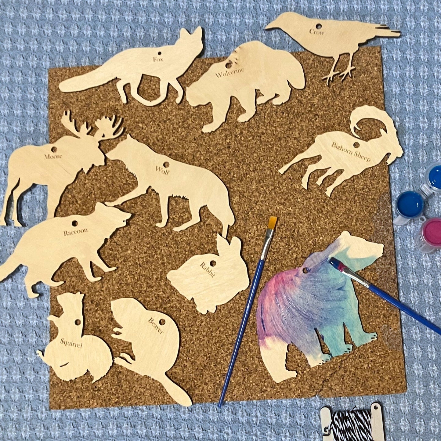 A photograph of 11 animal cutouts on a piece of cork. The bear has been painted with acrylic paints. There are 2 mini paint brushes and mini bobbin of string.