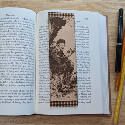 A photograph of the rectangular leather bookmark with the art of an elderly man reading engraved on it. The beareded man is sitting under a tree. The bookmark is resting on the pages of an open book.
