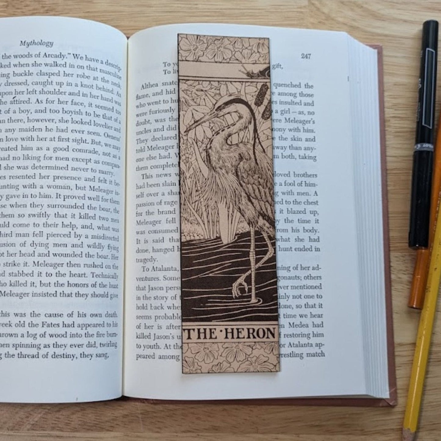 A rectangular leather bookmark with the ornate historical art of a heron engraved on it. The heron bookmark rests on the pages of an open book. Ornate Heron Leather Bookmark Bookmarks Raven King Crafts Ornate Heron Leather Bookmark Bookmarks Raven King Crafts