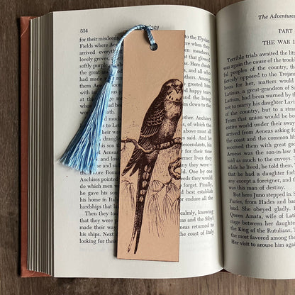 A rectangular bookmark with the art of a parakeet engraved on it. The bookmark has a blue tassel and is sitting on the pages of an open book.