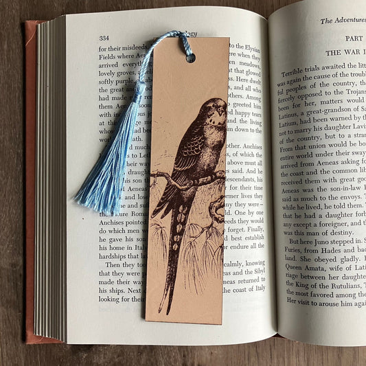 A rectangular bookmark with the art of a parakeet engraved on it. The bookmark has a blue tassel and is sitting on the pages of an open book. Parakeet Bookmark - Leather Bookmarks Raven King Crafts