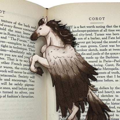 A close up of the leather pegasus bookmark. The bookmark is resting on the pages of an open book. Pegasus Bookmark - Leather Bookmarks Raven King Crafts