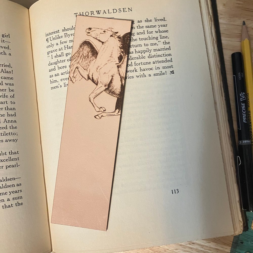 A leather bookmark with the art of a Pegasus engraved on it. The bookmark is resting on the pages of an open book. Pegasus Bookmark - Leather Bookmarks Raven King Crafts