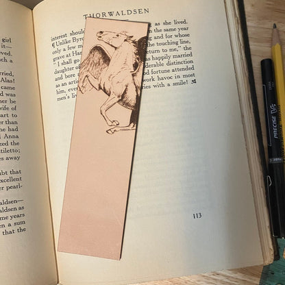 A leather bookmark with the art of a Pegasus engraved on it. The bookmark is resting on the pages of an open book. Pegasus Bookmark - Leather Bookmarks Raven King Crafts
