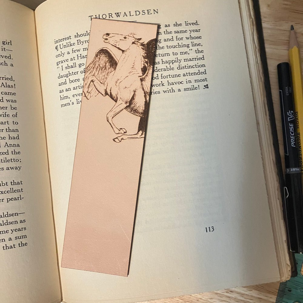 A leather bookmark with the art of a Pegasus engraved on it. The bookmark is resting on the pages of an open book.