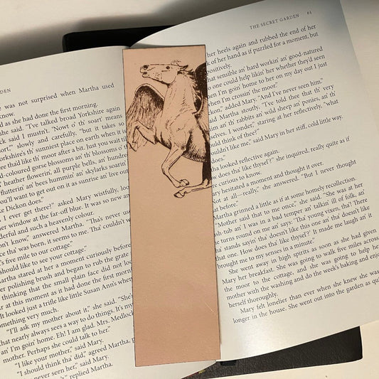 A leather bookmark with the art of a Pegasus engraved on it. The bookmark is resting on the pages of an open book.