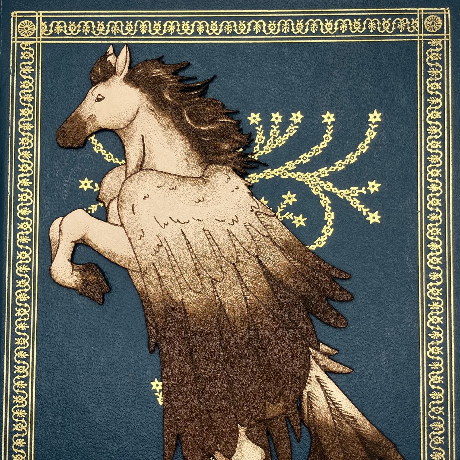 A detail shot of the leather pegasus bookmark. The bookmark is resting on the ornate blue and gold cover a of a book.