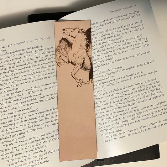 A leather bookmark with the art of a Pegasus engraved on it. The bookmark is resting on the pages of an open book. Pegasus Bookmark - Leather Bookmarks Raven King Crafts
