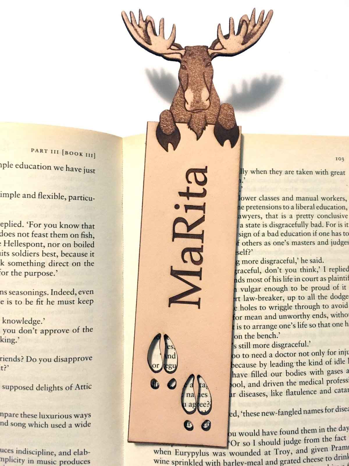 A photograph of exclusive personalizable moose leather bookmark resting on the pages of an open book. Personalizable Moose Leather Bookmark - Unique Gift for Book Lovers Bookmarks Raven King Crafts