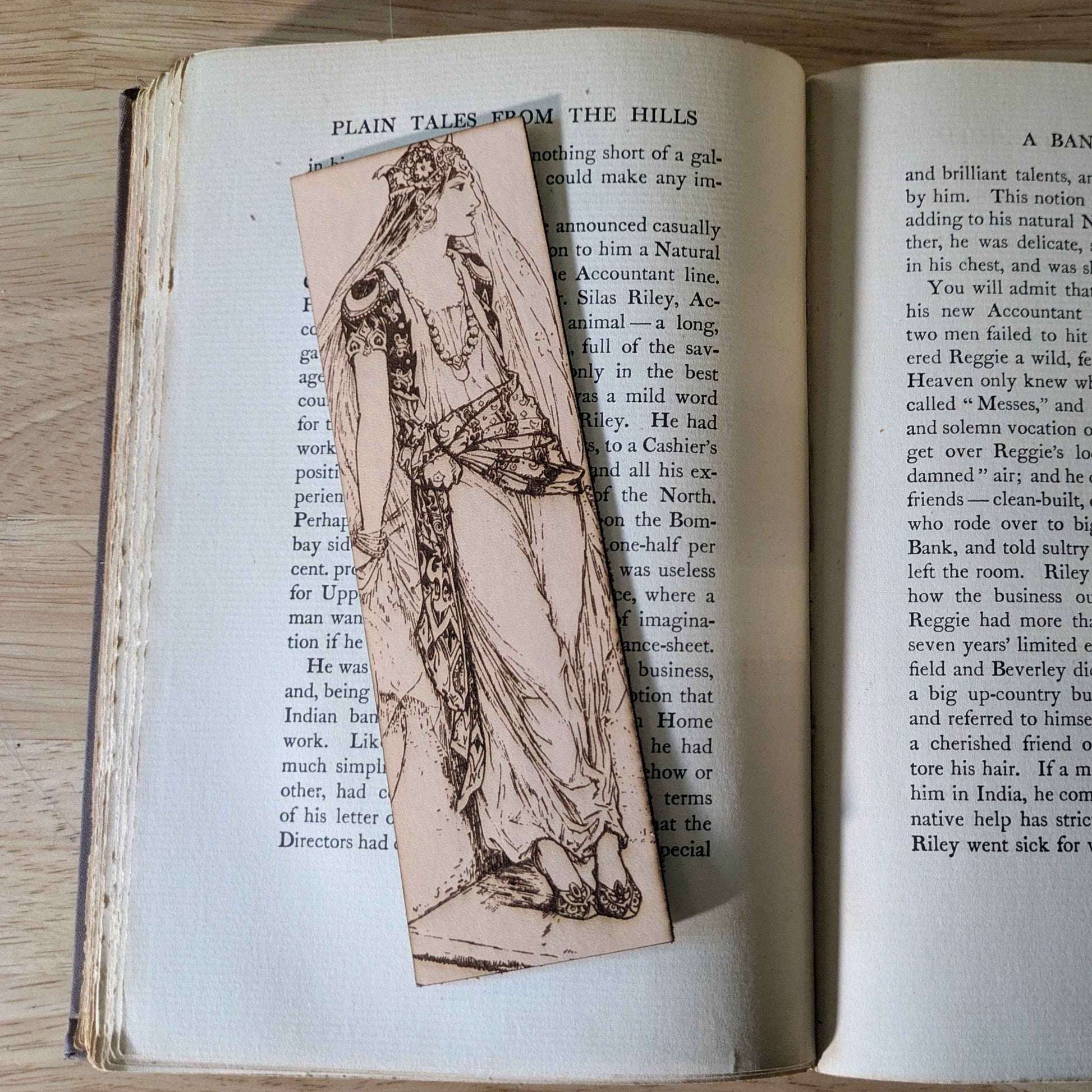 A leather rectangular bookmark with the antique art of a princess waiting engraved on it. The leather bookmark is resting on the pages of an open book. Princess Waiting - Leather Bookmark Bookmarks Raven King Crafts