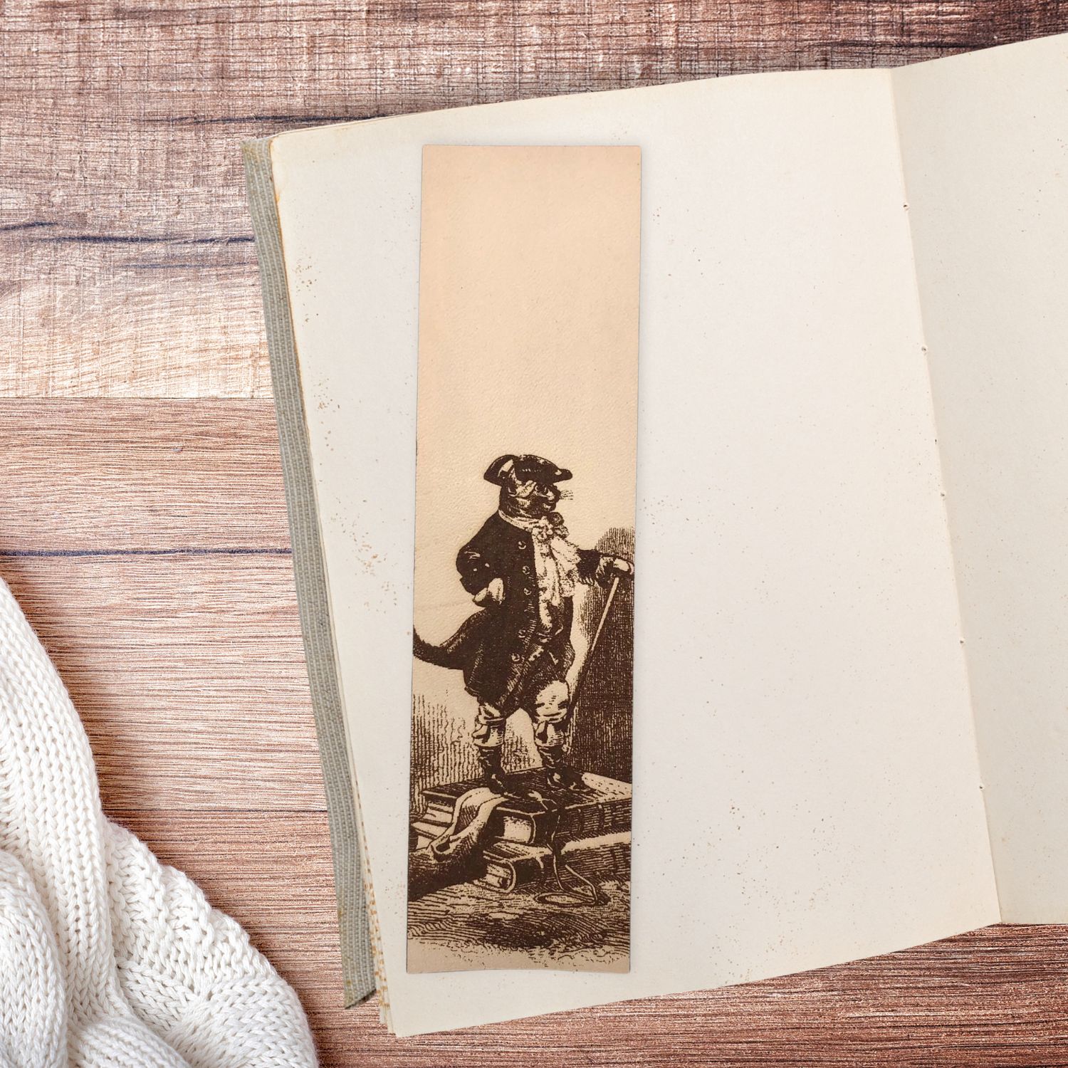 A rectangular bookmark with the historical art of puss in boots engraved on it. The bookmark is sitting on the pages of an open book.