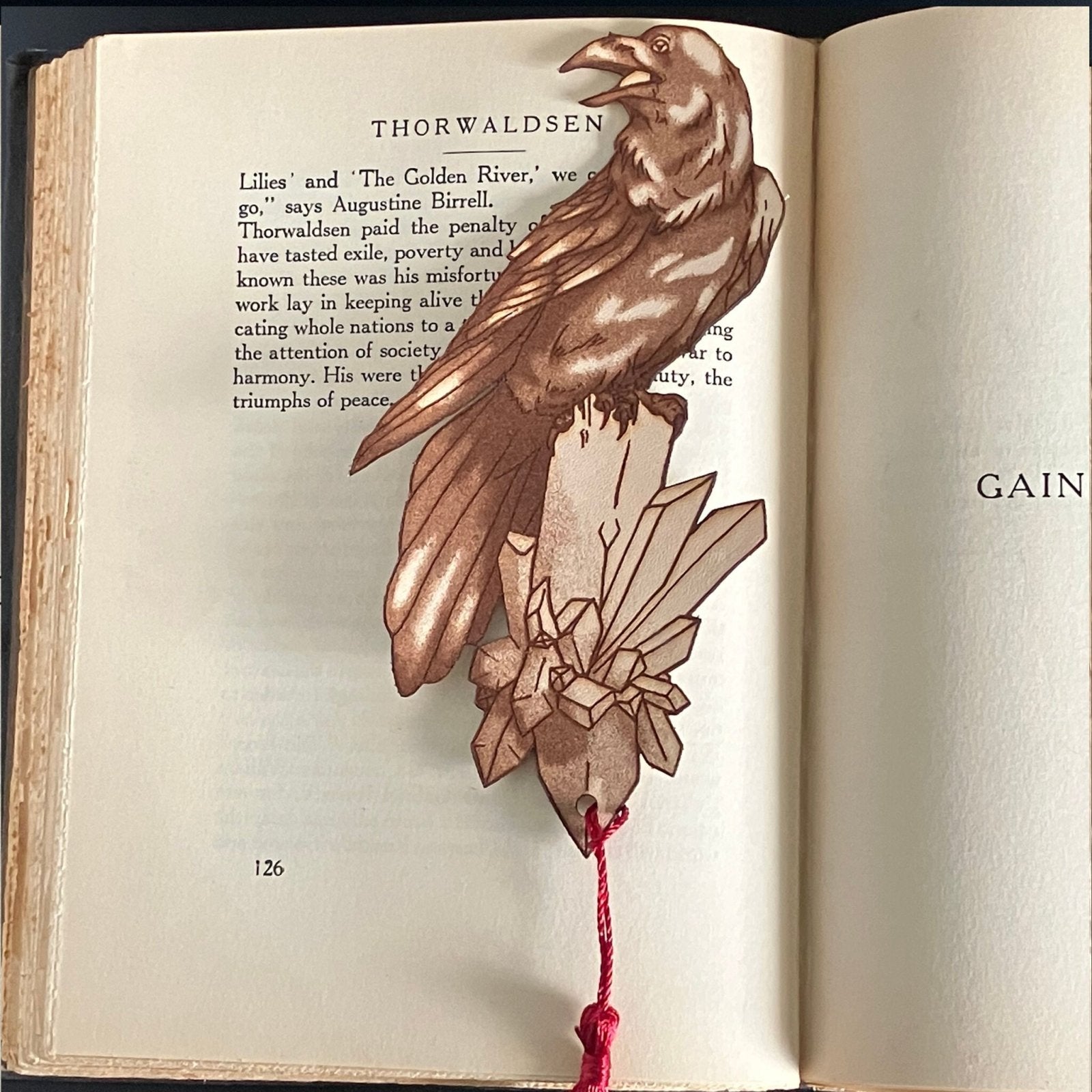 A raven shaped bookmark with a red tassel. The bookmark is resting on the pages of an open book.