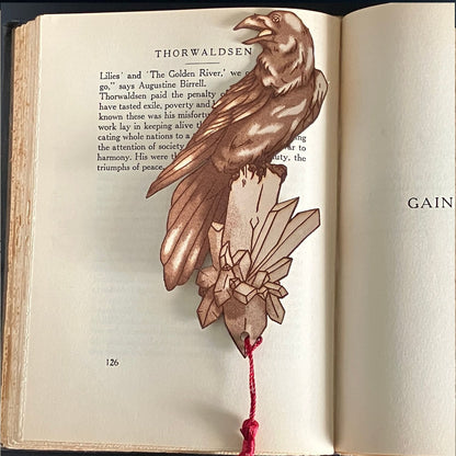 A raven shaped bookmark with a red tassel. The bookmark is resting on the pages of an open book.