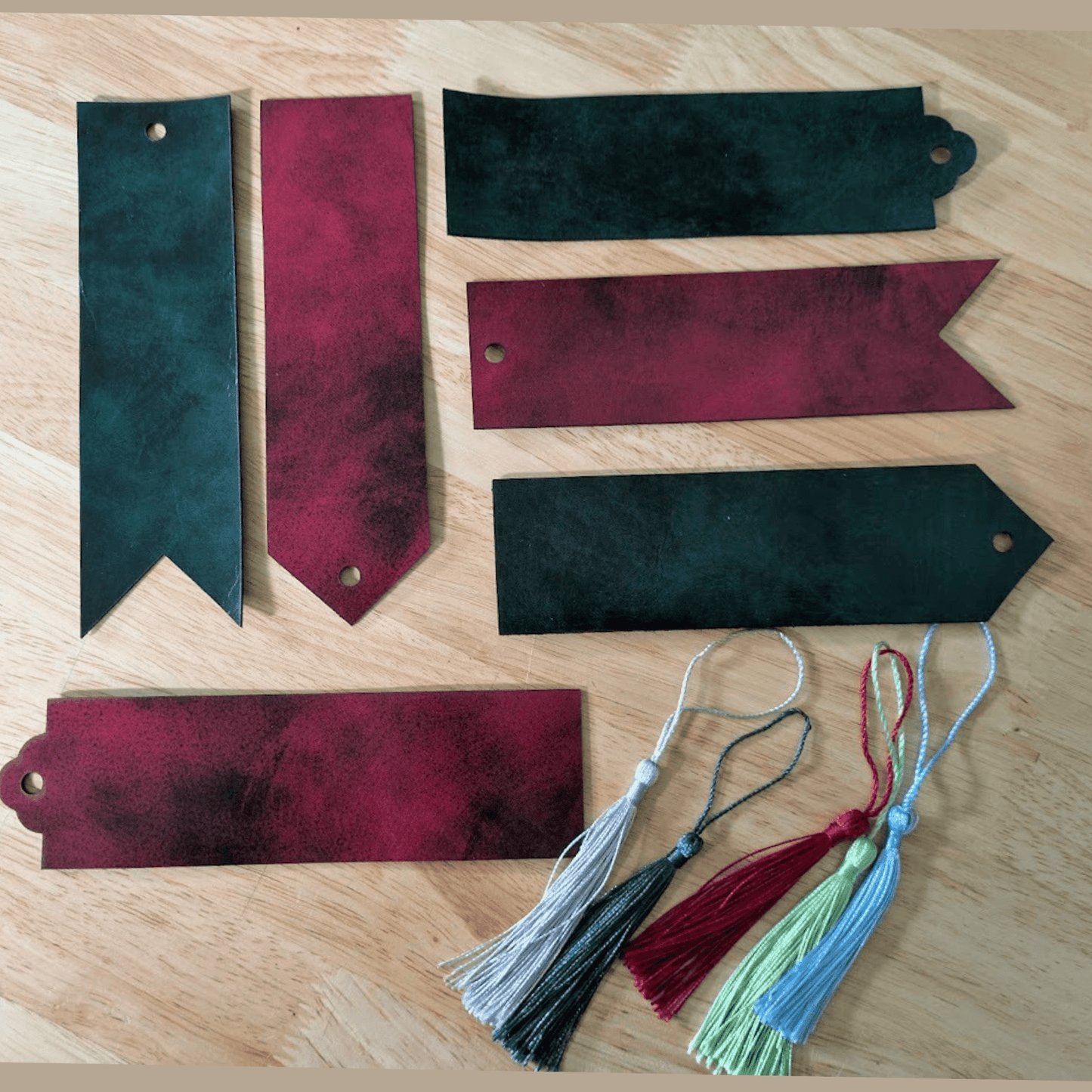 A photograph of the green and red black dyed leather bookmarks. Top left is ribbon style green and black, red and black pointed, green rounded, red ribbon, green pointed, red rounded and the tassels. Red or Green Leather Bookmarks with Tassel Bookmarks Raven King Crafts Red or Green Leather Bookmarks with Tassel Bookmarks Raven King Crafts