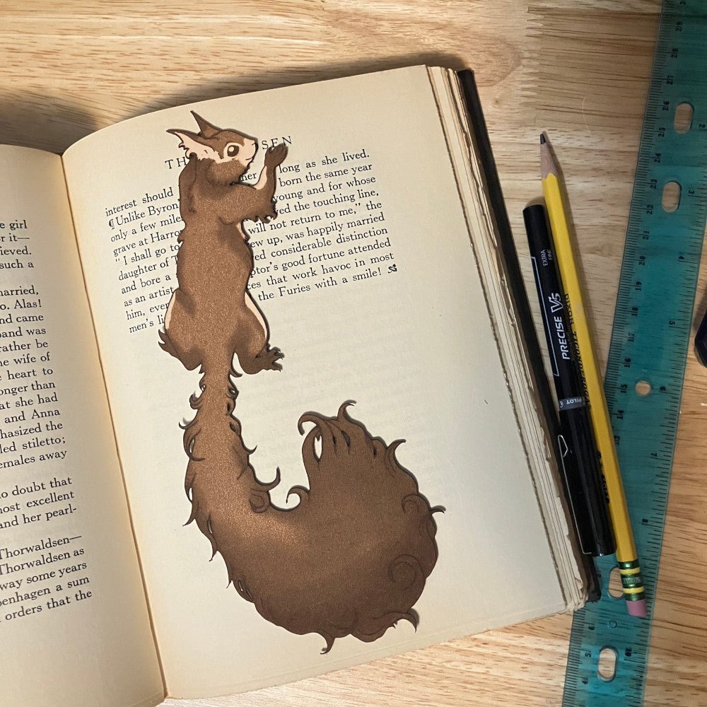 This photograph shows a red squirrel shaped bookmark resting on top of the pages of an open book. Next to the book are a pen, pencil and a ruler for scale.