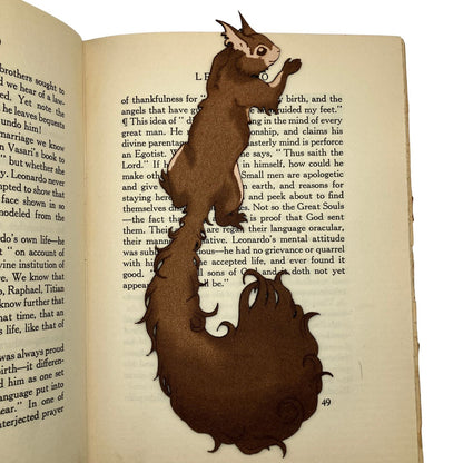 A photo of the red squirrel bookmark resting on the pages of an open book. Red Squirrel Bookmark - Leather Bookmarks Raven King Crafts