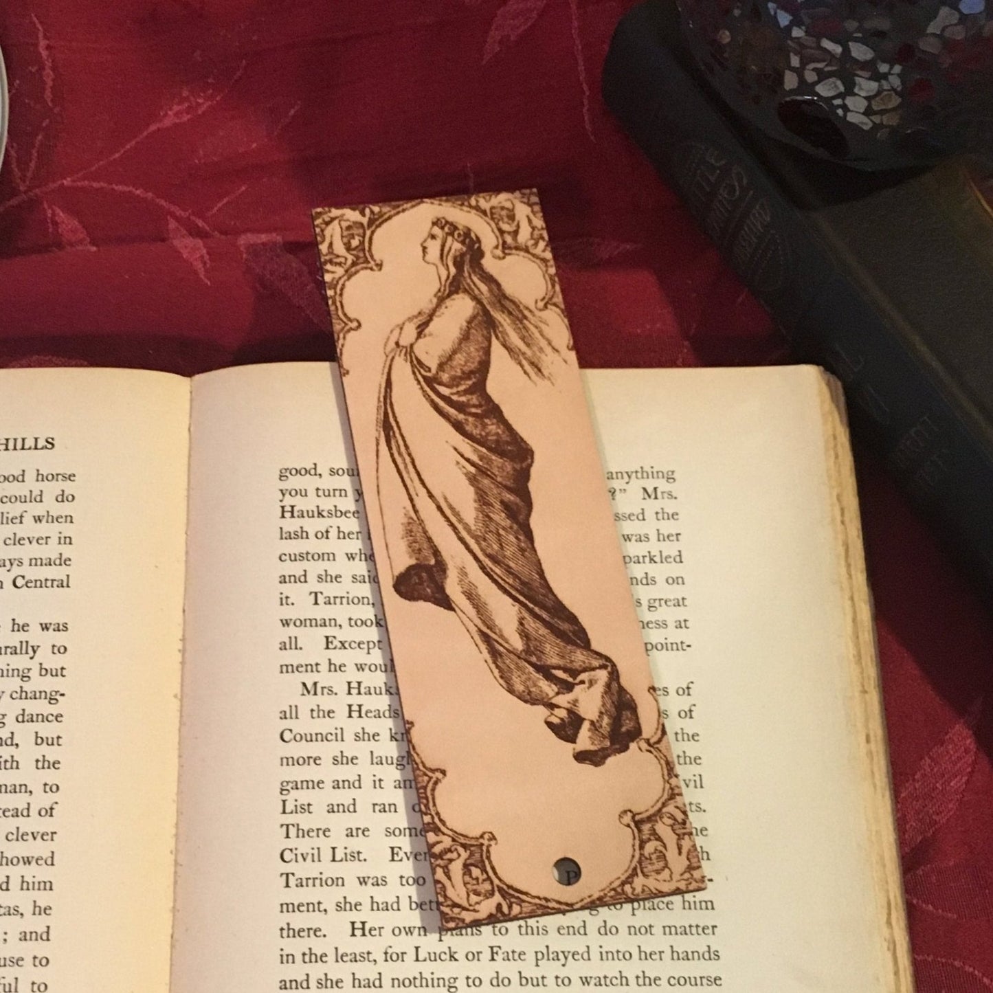 The bookmark of a robed woman with a crown with an ornate framing. The bookmark is resting on a book.