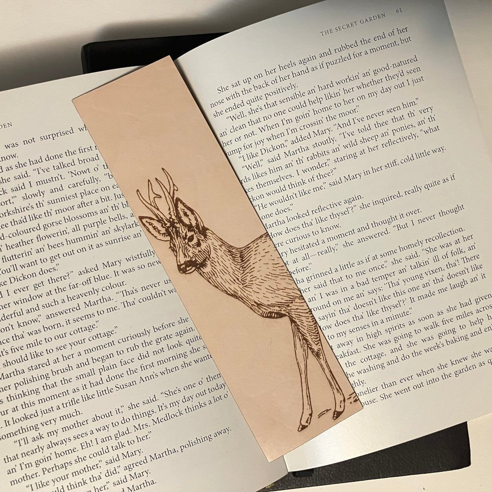 The photograph shows a leather bookmark with the art of a roe deer on it. The bookmark is resting on the pages of an open book.