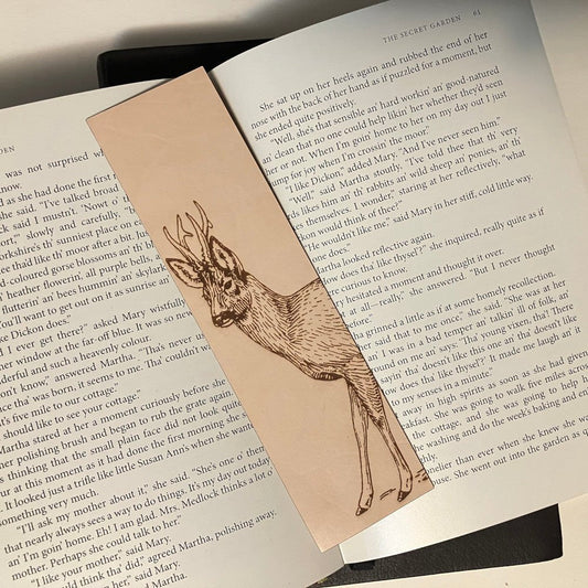 The photograph shows a leather bookmark with the art of a roe deer on it. The bookmark is resting on the pages of an open book. Roe Deer Bookmark - Leather Bookmarks Raven King Crafts