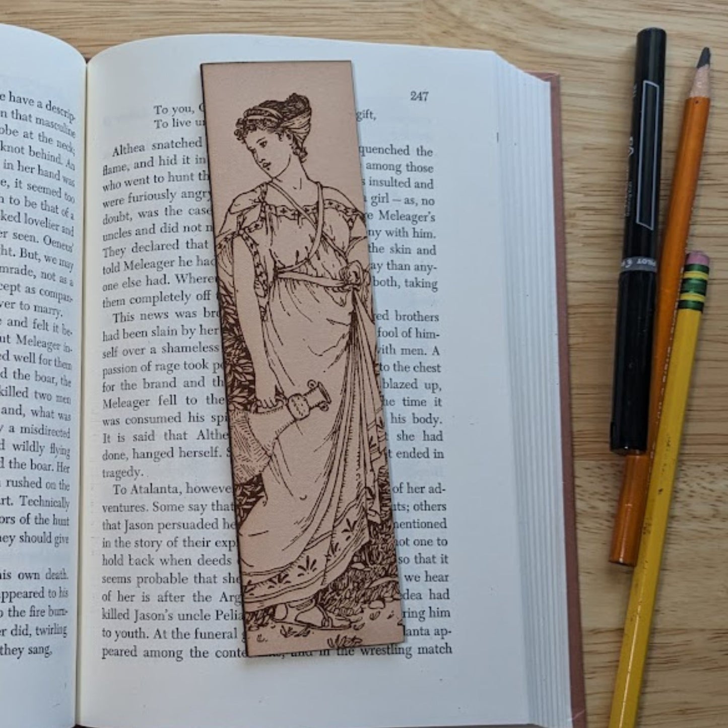 A rectangular leather bookmark with the antique art of a Roman or Greek woman. The woman is wearing sandals and a toga. She also has her hair tied up. The bookmark is resting on the pages of an open book.