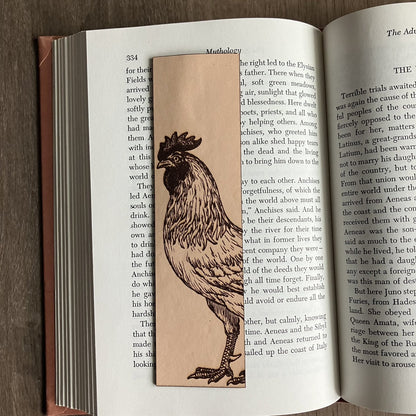 A rectangular bookmark with the art of a rooster engraved on it. The bookmark is resting on the pages of an open book.