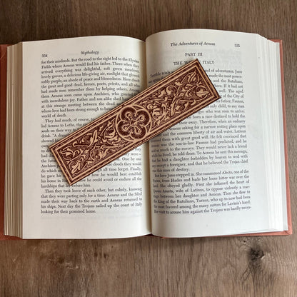 Photograph of a leather bookmark with a decorative design engraved on it. At the center of the design is a rounded cross design. The bookmark is resting on the pages of an open book.