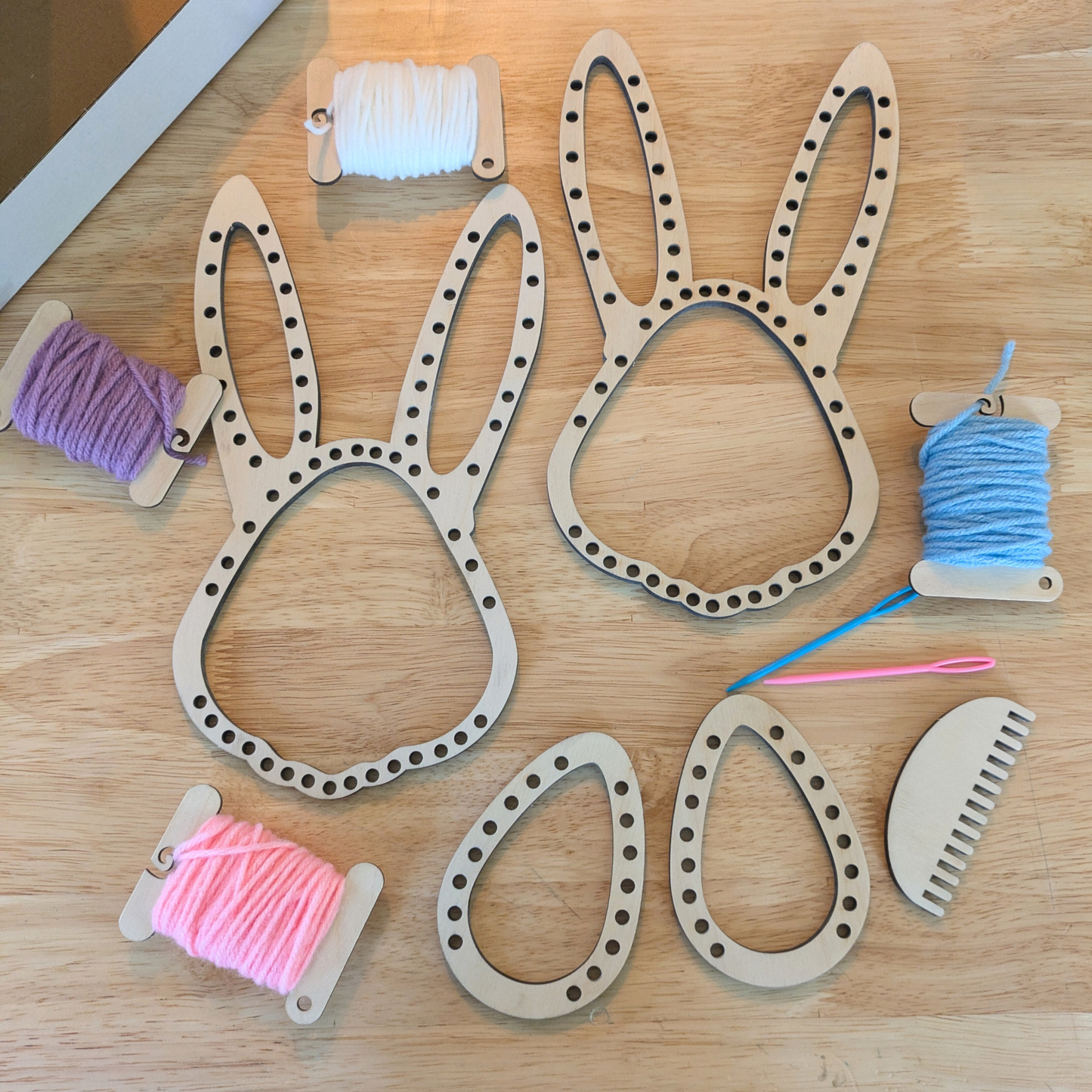 A photograph of the Easter Loom set, including 2 rabbit head shaped looms, 2 egg shaped loos, a comb, 2 needles and 4 spools of spring colored yarn.