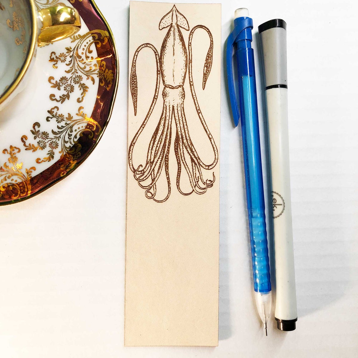 The squid bookmarker next to a pencil and pen for scale. There is also a tea cup to the right.