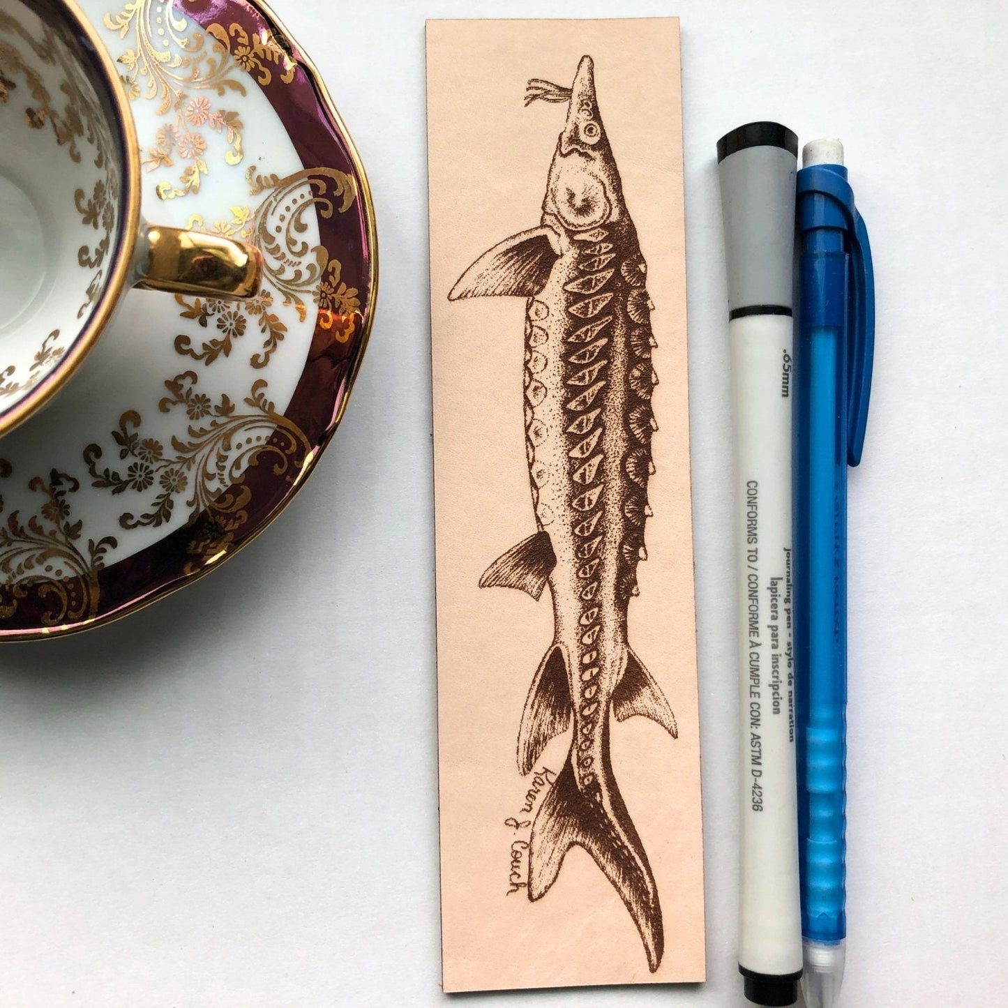 A fish engraved on a leather bookmark. Next to it is a pen and mechanical pencil for scale and on the right tis a tea cup. Sturgeon Bookmark Bookmarks Raven King Crafts