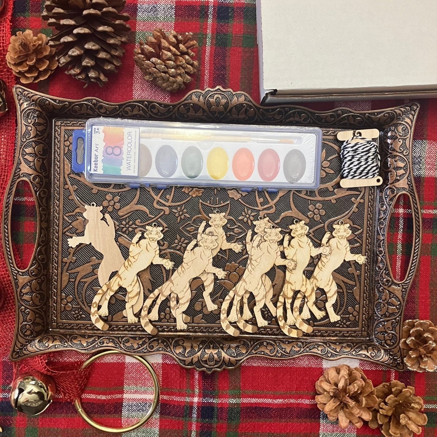 A metal tray holding 8 wooden tiger ornaments. This kit option also shows watercolors and string. Tiger Shaped Wooden Ornaments Art & Craft Kits Raven King Crafts