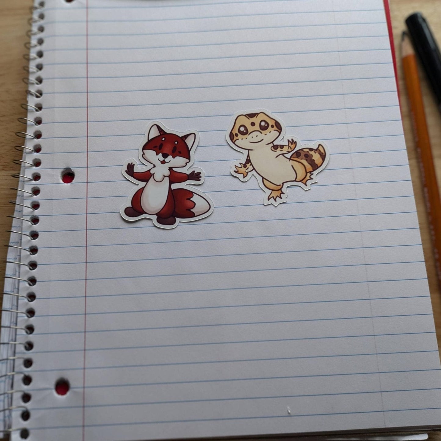 A photograph of the chibi fox (left) and the chibi gecko (right) resting on the page of an open notebook. Vinyl Sticker Packs - Pick Your Stickers! stickers Raven King Crafts Vinyl Sticker Packs - Pick Your Stickers! stickers Raven King Crafts