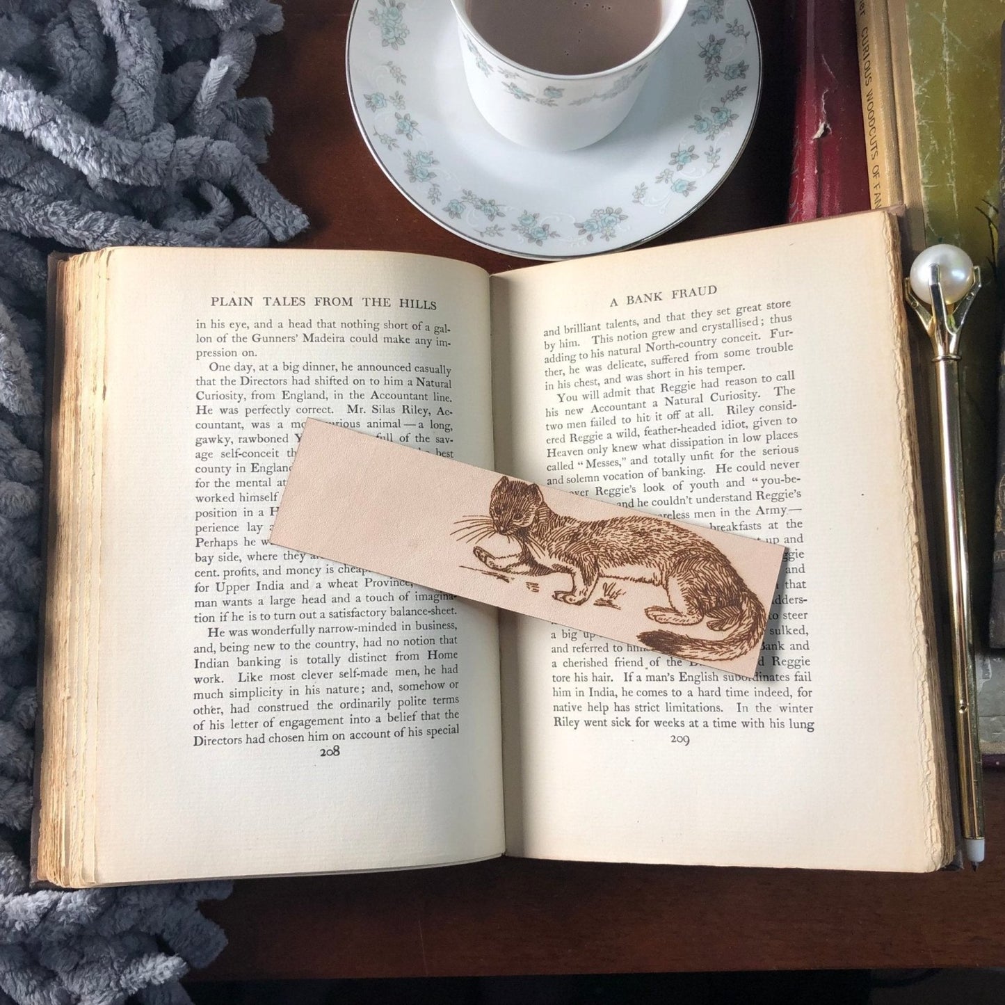 A leather bookmark of a polecat on an antique book. Next to the book is a pen, and a tea cup.