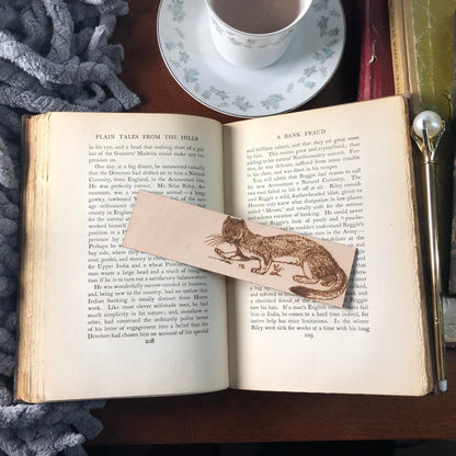 A leather bookmark of a polecat on an antique book. Next to the book is a pen, and a tea cup. Weasel Bookmark Bookmarks Raven King Crafts