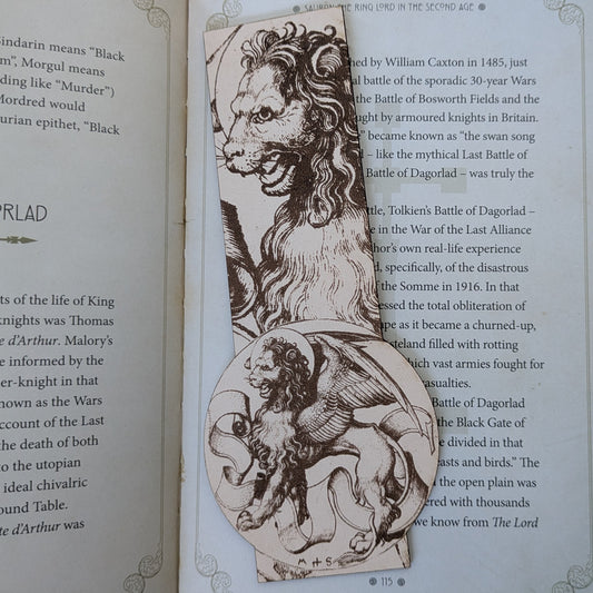 A leather bookmark with the art of illustration of a winged lion engraved on it. The bookmark is resting on the pages of an open book.