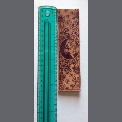 The bookmark of a Greek or Roman woman and wildflowers next to a ruler for scale Woman and Gust of Flowers Leather Bookmark Bookmarks Raven King Crafts
